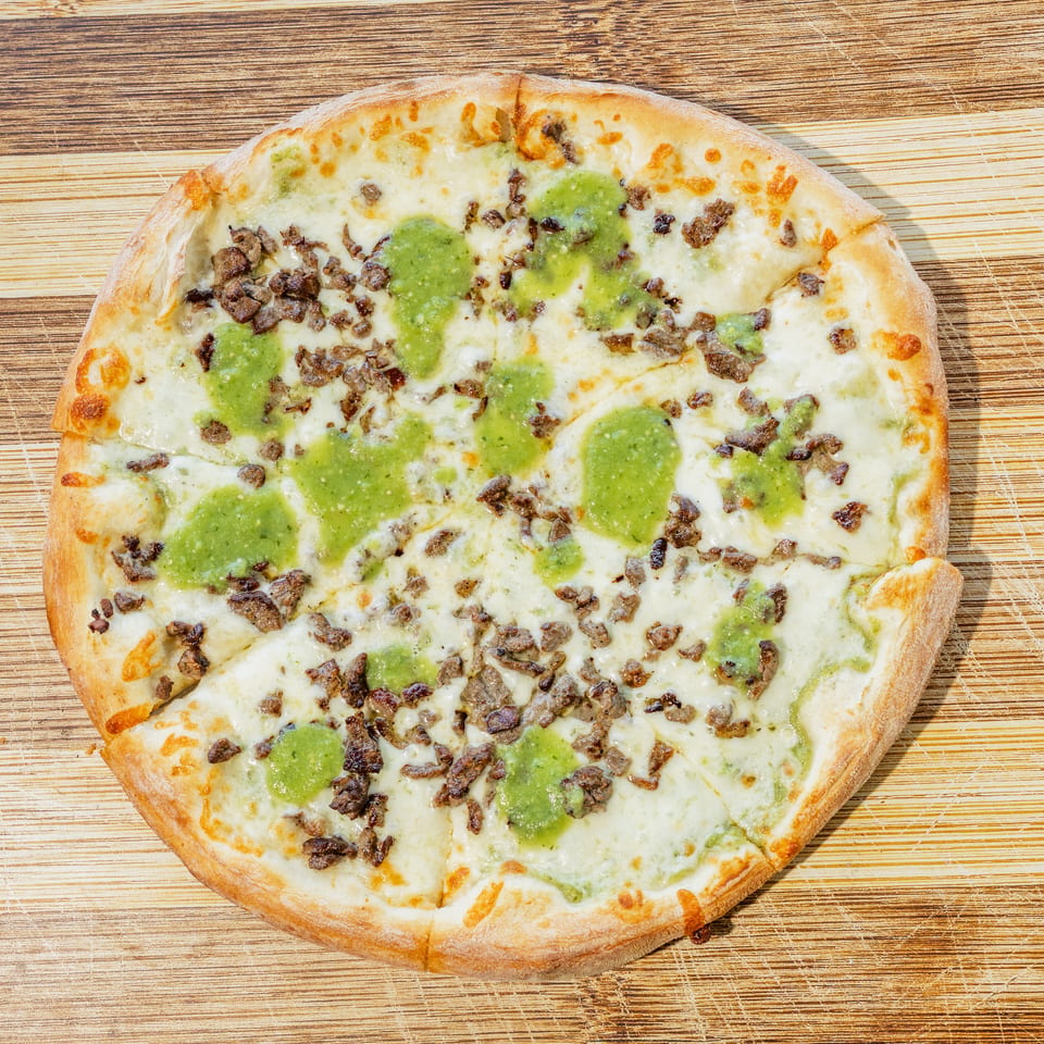 Round cheesy pizza topped with browned meat pieces and green sauce on a rustic wooden tabletop.