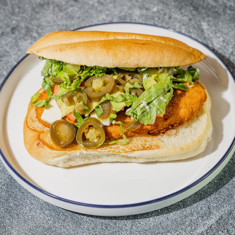 Crispy breaded sandwich topped with lettuce, tomatoes, cheese, avocado, and sliced jalapeños on a white plate.