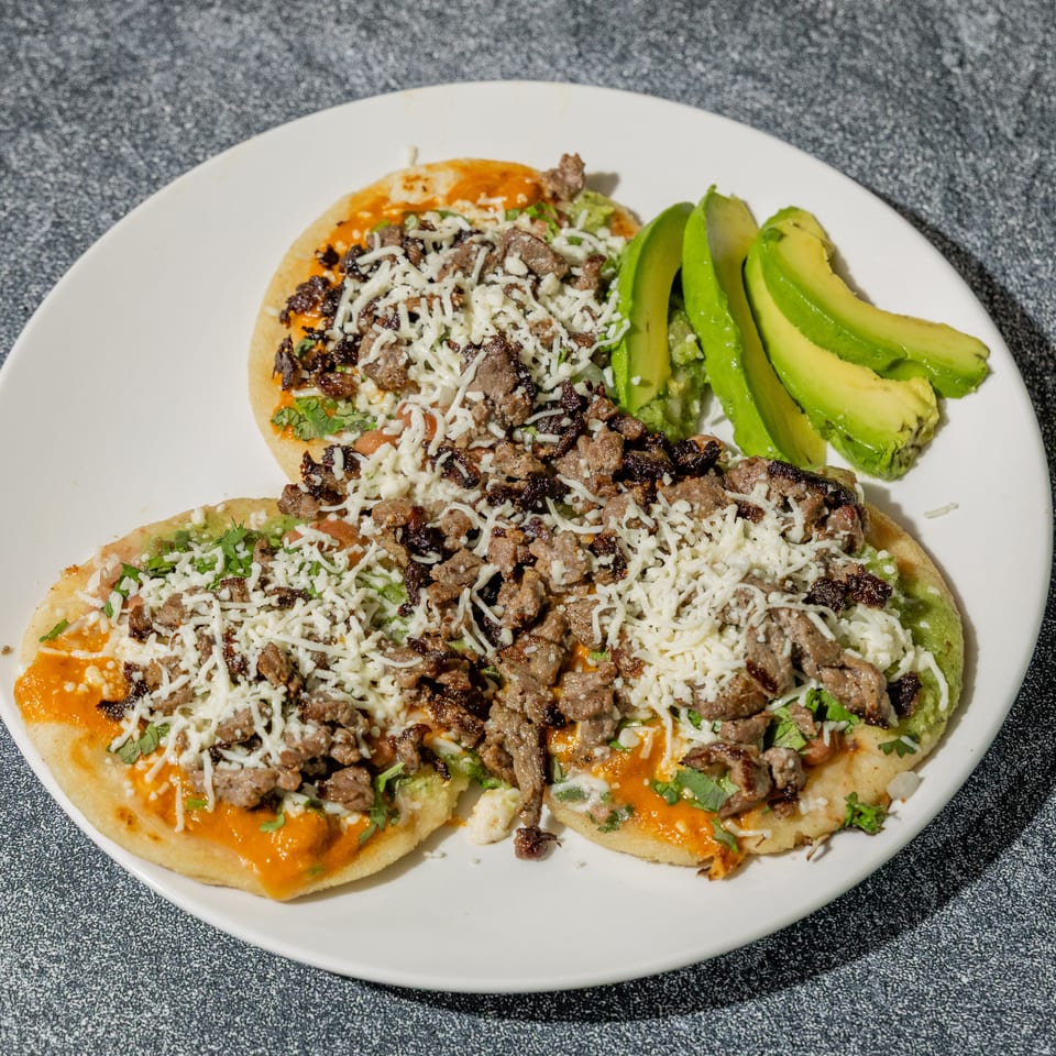 Two meat-topped, cheese-covered tostadas with beans, salsa, cilantro, and avocado slices on a white plate.