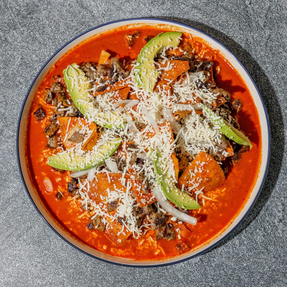 Plate of red-sauced chilaquiles topped with grilled meat, avocado slices, onions, and crumbled cheese.